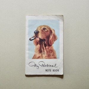 May include: A rectangular notebook with a white cover featuring a centered illustration of a golden-brown dog holding a dark leash in its mouth against a light blue background. The text "My Personal NOTE BOOK" is printed in black below the dog.
