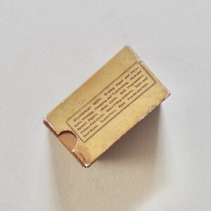 Pode incluir: Uma caixa de papelaria vintage com uma apar&ecirc;ncia desgastada e envelhecida. A caixa &eacute; retangular com uma aba e apresenta texto impresso indicando que &eacute; do "Stationery Dept." e lista v&aacute;rios artigos de papelaria. A caixa &eacute; de cor castanho claro.