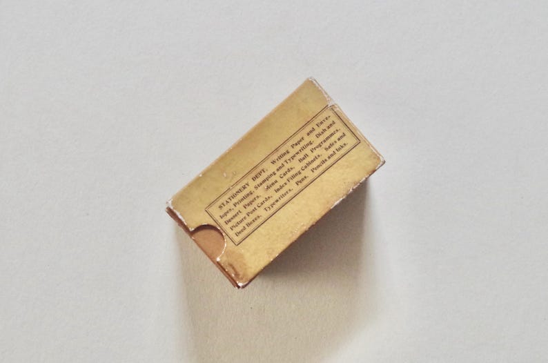 Pode incluir: Uma caixa de papelaria vintage com uma apar&ecirc;ncia desgastada e envelhecida. A caixa &eacute; retangular com uma aba e apresenta texto impresso indicando que &eacute; do "Stationery Dept." e lista v&aacute;rios artigos de papelaria. A caixa &eacute; de cor castanho claro.