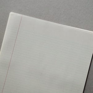 May include: A white, lined notebook with a red vertical margin line. The notebook is open, revealing the ruled pages. The background is a solid gray color. The notebook is likely for taking notes or writing.