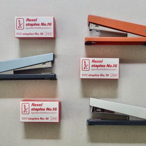 May include: A collection of staplers in orange, pale blue, and white, with boxes of Rexel staples No. 16. The boxes are red and white, with text indicating 1000 staples per box. The staplers are displayed on a neutral background.