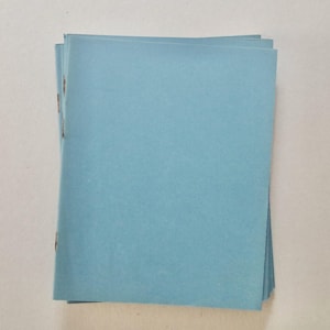 May include: A pile of light blue paper booklets, stapled along the left edge. The booklets have a smooth texture and are neatly stacked. The background is a plain, off-white surface.