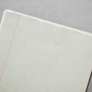 Op de afbeelding: Een stapel gelijnd papier met een rode verticale margelijn. Het papier is wit met horizontale lijnen en de randen van de stapel zijn zichtbaar. De achtergrond is een lichtgrijze ondergrond.
