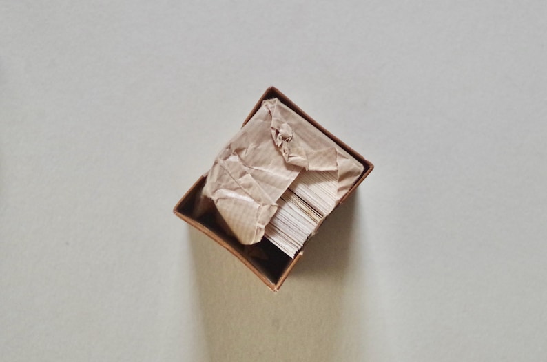 Pode incluir: Uma pequena caixa retangular aberta com exterior castanho. No interior, uma pilha de cart&otilde;es brancos &eacute; parcialmente vis&iacute;vel, coberta por papel castanho amassado. A caixa est&aacute; sobre uma superf&iacute;cie cinza claro.