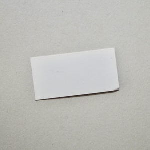 Pode incluir: Um pequeno cart&atilde;o retangular branco com um canto ligeiramente dobrado, colocado sobre uma superf&iacute;cie cinza claro. O cart&atilde;o &eacute; liso, sem texto ou design vis&iacute;vel, e parece ser feito de papel ou cart&atilde;o.