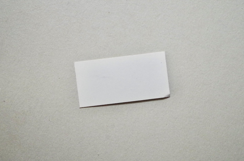 Pode incluir: Um pequeno cart&atilde;o retangular branco com um canto ligeiramente dobrado, colocado sobre uma superf&iacute;cie cinza claro. O cart&atilde;o &eacute; liso, sem texto ou design vis&iacute;vel, e parece ser feito de papel ou cart&atilde;o.