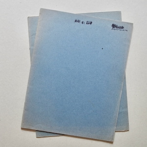 May include: Two pale blue booklets are stacked on a white surface. Each booklet has handwritten text in black ink at the top. The paper has a slightly textured appearance. The overall composition is simple and clean.