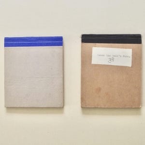 Könnte beinhalten: Zwei rechteckige Notizblöcke. Der linke Block hat einen hellgrauen Einband mit einem blauen Streifen am oberen Rand. Der rechte Block hat einen beigen Einband mit einem schwarzen Rand und einem weißen Etikett "Guess the Doll's Name. 3d".