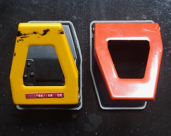 May include: Two vintage hole punches, one yellow and one red, sit side-by-side on a dark surface. The yellow punch has a label with the numbers "1768/139 02". Both punches have a rectangular shape with a large opening.