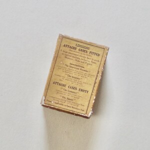 Pode incluir: Uma caixa retangular vintage com texto, provavelmente para maletas. A caixa tem um r&oacute;tulo dourado com texto preto, incluindo "Attache Cases Fitted" e "Attache Cases Empty". A caixa parece envelhecida e desgastada.