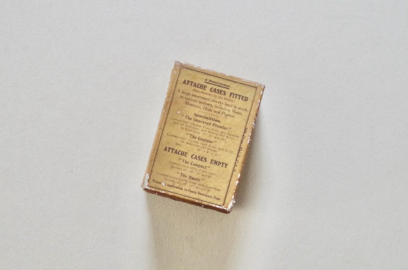 Pode incluir: Uma caixa retangular vintage com texto, provavelmente para maletas. A caixa tem um r&oacute;tulo dourado com texto preto, incluindo "Attache Cases Fitted" e "Attache Cases Empty". A caixa parece envelhecida e desgastada.