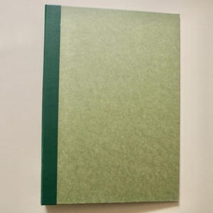 Puede incluir: Un cuaderno rectangular con una cubierta de color verde salvia claro y una franja vertical en el lomo de color verde bosque oscuro sólido. La cubierta tiene una textura sutilmente moteada y la encuadernación es recta en el borde izquierdo.