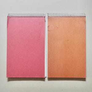 May include: Two rectangular spiral-bound notebooks placed side by side. The notebook on the left has a solid dark pink cover, while the notebook on the right has a solid light brown cover with visible circular marks on the upper surface.