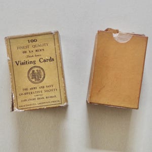 Pode incluir: Duas caixas vintage de cart&otilde;es de visita. Uma caixa est&aacute; fechada e a outra est&aacute; aberta. A caixa fechada tem o texto "100 Finest Quality Visiting Cards" e o nome "De La Rue's". As caixas s&atilde;o castanhas e t&ecirc;m um aspeto desgastado.