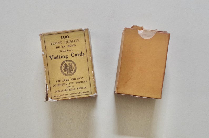 Pode incluir: Duas caixas vintage de cart&otilde;es de visita. Uma caixa est&aacute; fechada e a outra est&aacute; aberta. A caixa fechada tem o texto "100 Finest Quality Visiting Cards" e o nome "De La Rue's". As caixas s&atilde;o castanhas e t&ecirc;m um aspeto desgastado.