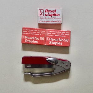 May include: An overhead shot of office supplies, including a red and silver stapler and three boxes of Rexel staples. The boxes are red and white, with the text "Rexel No 56 Staples" and "1000 staples No.56266".