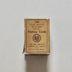 Pode incluir: Uma caixa retangular de "Visiting Cards" bege com texto preto, incluindo "Finest Quality" e "De La Rue's". A caixa, ligeiramente desgastada, tamb&eacute;m mostra o nome "The Army and Navy Co-operative Society".
