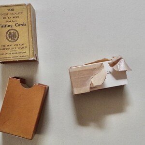 Pode incluir: Uma vista a&eacute;rea de tr&ecirc;s caixas de cart&otilde;es vintage sobre uma superf&iacute;cie clara. Uma caixa intacta com a inscri&ccedil;&atilde;o "100 FINEST QUALITY Visiting Cards". As outras duas caixas est&atilde;o parcialmente abertas ou danificadas. As caixas s&atilde;o bege e castanhas.