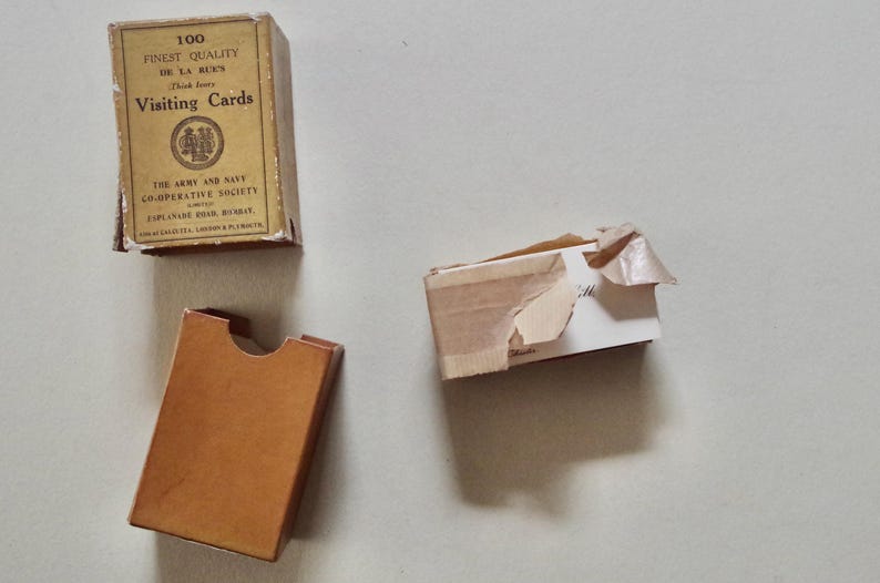 Pode incluir: Uma vista a&eacute;rea de tr&ecirc;s caixas de cart&otilde;es vintage sobre uma superf&iacute;cie clara. Uma caixa intacta com a inscri&ccedil;&atilde;o "100 FINEST QUALITY Visiting Cards". As outras duas caixas est&atilde;o parcialmente abertas ou danificadas. As caixas s&atilde;o bege e castanhas.