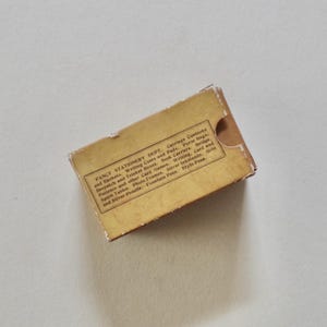 Pode incluir: Uma pequena caixa de papelaria vintage retangular em tons de bege e castanho. A caixa tem um aspeto desgastado e apresenta texto impresso, incluindo "Fancy Stationery Dept." e uma lista de artigos. A caixa est&aacute; ligeiramente aberta.