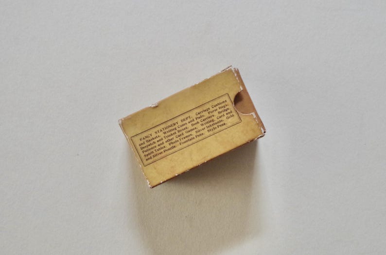 Pode incluir: Uma pequena caixa de papelaria vintage retangular em tons de bege e castanho. A caixa tem um aspeto desgastado e apresenta texto impresso, incluindo "Fancy Stationery Dept." e uma lista de artigos. A caixa est&aacute; ligeiramente aberta.