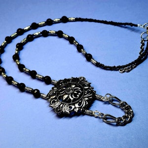 May include: A black and silver beaded necklace with a large, ornate silver pendant. The pendant features a floral design.