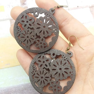 Pode incluir: Brincos de madeira castanha escura com um design floral recortado. Os brincos s&atilde;o circulares com intrincados padr&otilde;es florais e ganchos de orelha de cor bronze antigo. Os brincos s&atilde;o segurados em uma m&atilde;o.