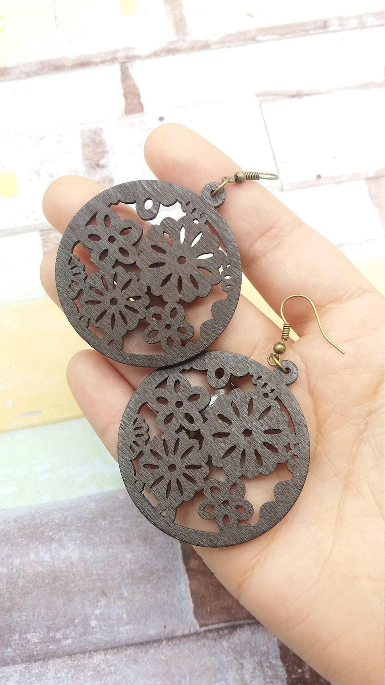 Pode incluir: Brincos de madeira castanha escura com um design floral recortado. Os brincos s&atilde;o circulares com intrincados padr&otilde;es florais e ganchos de orelha de cor bronze antigo. Os brincos s&atilde;o segurados em uma m&atilde;o.