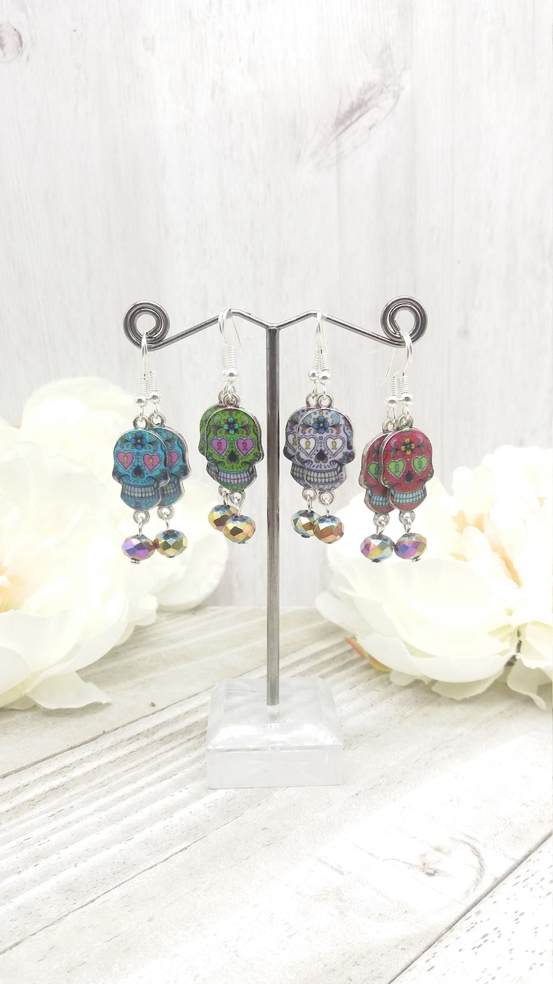 May include: Four pairs of dangle earrings featuring sugar skull designs with colorful floral patterns. Each pair has two crystal beads hanging below the skull charm.