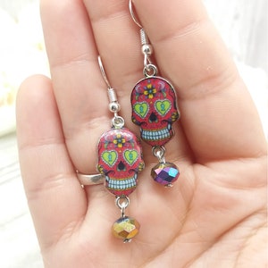 May include: A pair of silver dangle earrings featuring red, white, and blue sugar skull charms with green accents and iridescent glass beads.
