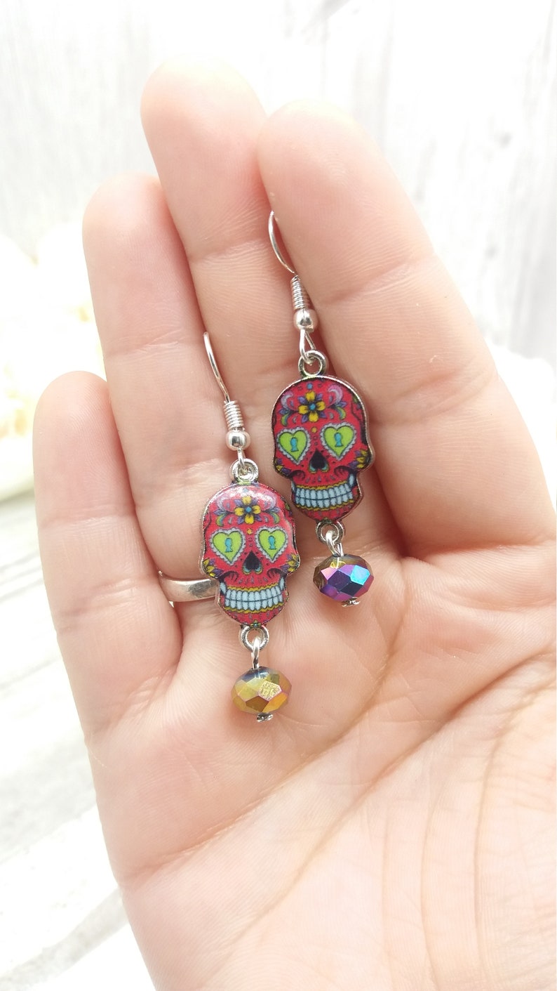 May include: A pair of silver dangle earrings featuring red, white, and blue sugar skull charms with green accents and iridescent glass beads.