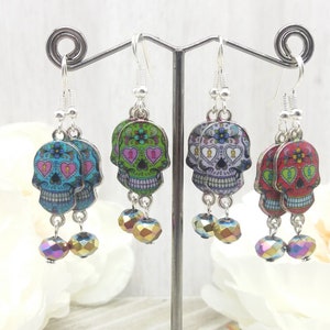 May include: Three pairs of silver dangle earrings with colorful sugar skull charms and iridescent crystal beads. The sugar skulls have pink, green, and red designs with black outlines.