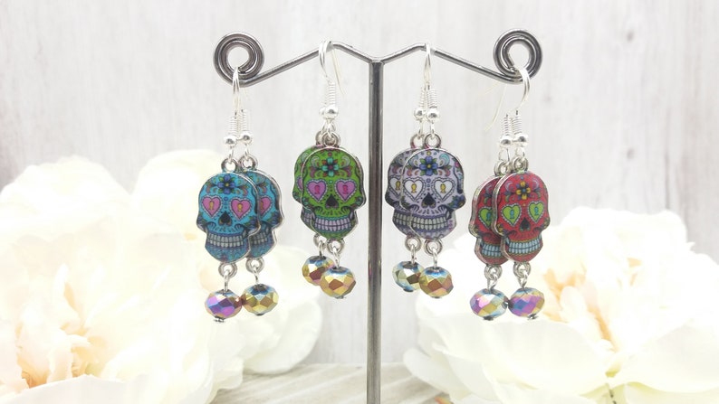 May include: Three pairs of silver dangle earrings with colorful sugar skull charms and iridescent crystal beads. The sugar skulls have pink, green, and red designs with black outlines.