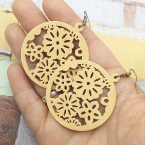 Pode incluir: Um par de brincos de madeira castanha clara. Cada brinco tem formato circular com um design floral vazado. Os brincos s&atilde;o segurados em uma m&atilde;o, mostrando os detalhes intrincados e os tons quentes da madeira.