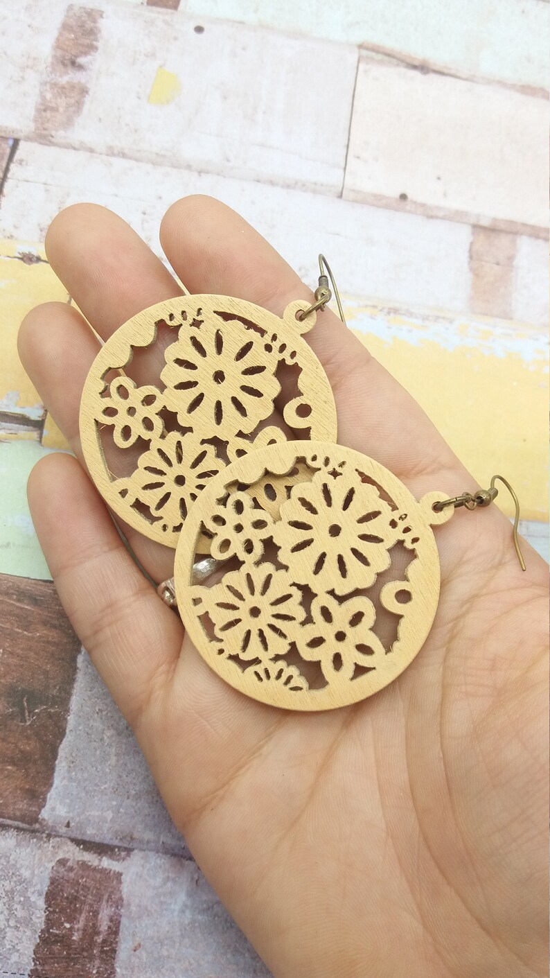 Pode incluir: Um par de brincos de madeira castanha clara. Cada brinco tem formato circular com um design floral vazado. Os brincos s&atilde;o segurados em uma m&atilde;o, mostrando os detalhes intrincados e os tons quentes da madeira.