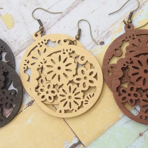 Pode incluir: Tr&ecirc;s pares de brincos de madeira em tons variados de castanho e bege. Cada brinco apresenta um design circular com intrincados recortes florais. Os brincos penduram-se em fios de orelha cor de bronze, mostrando uma est&eacute;tica natural e artesanal.