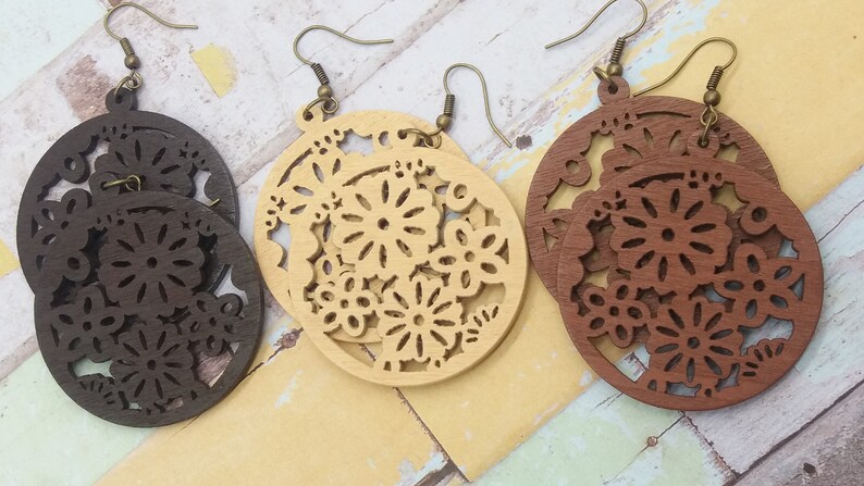 Pode incluir: Tr&ecirc;s pares de brincos de madeira em tons variados de castanho e bege. Cada brinco apresenta um design circular com intrincados recortes florais. Os brincos penduram-se em fios de orelha cor de bronze, mostrando uma est&eacute;tica natural e artesanal.