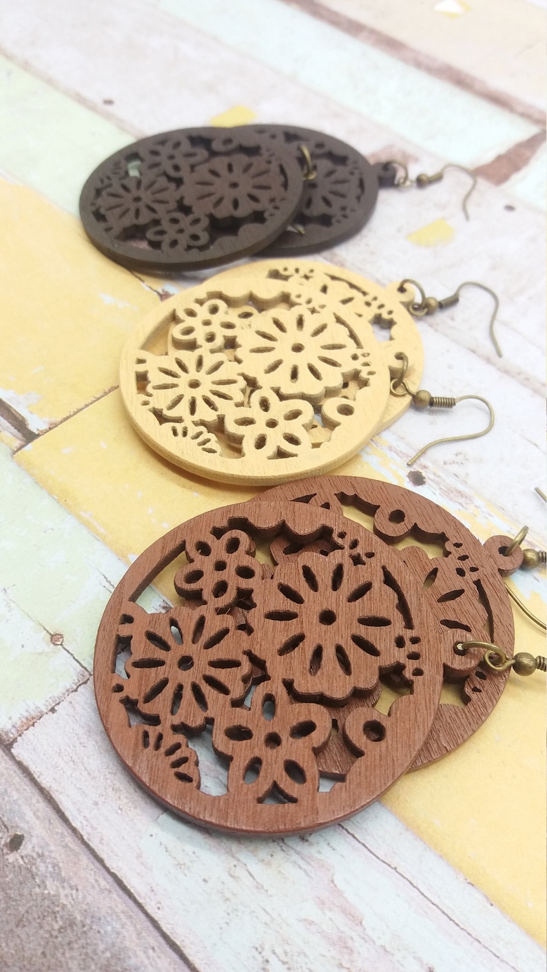 Pode incluir: Brincos de madeira em tr&ecirc;s cores: castanho escuro, castanho claro e bege. Cada brinco tem formato circular com um design floral vazado. Os brincos t&ecirc;m ganchos de orelha na cor bronze.
