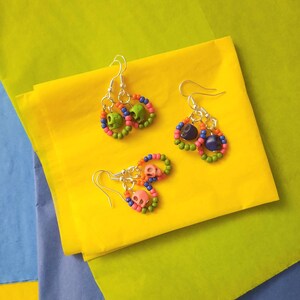 K&ouml;nnte beinhalten: Ein Paar Ohrringe mit gr&uuml;nen, blauen und rosa Perlen und Totenkopf-Anh&auml;ngern. Die Ohrringe liegen auf einer gelben Oberfl&auml;che.
