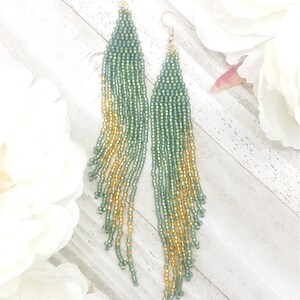 May include: A pair of long, green and gold beaded earrings. The earrings are made with small, round beads that are strung together in a cascading pattern. The beads are a gradient of green and gold, with the green beads at the top and the gold beads at the bottom.