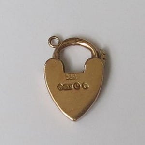 May include: A gold heart-shaped padlock charm featuring a small circular jump ring at the top. The surface of the charm displays engraved hallmarks, including the letters JBL and the number 375, along with several small, distinct geometric symbols.