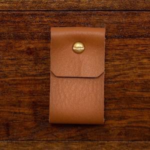 May include: A close-up of a wooden drawer with a small, tan leather pull. The drawer is part of a larger wooden piece, with a white cylindrical planter holding a green plant in the background. The leather pull is secured with a gold-colored stud.
