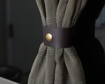 May include: A gray kitchen towel with fringe hangs from a stainless steel oven handle. A dark brown leather strap with a gold button secures the towel. The oven is next to white cabinets with black hardware.