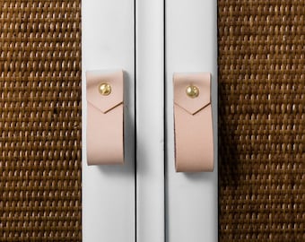 May include: White cabinet with woven brown door panels and pale pink leather drawer pulls and door handles. The drawer pulls are long and rectangular, secured with gold-colored rivets. The door handles are short and square, also secured with gold rivets.