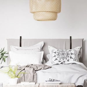 May include: A white bed with a grey headboard and white bedding. The bed has a woven wicker pendant light hanging above it. There are two small wooden stools in front of the bed and a potted cactus plant in the corner of the room.