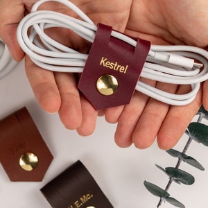 May include: Six leather cord organizers with gold metal accents and personalized names. The names are "Kestrel", "Addison", "Travis", "Stephanie", "N.E.Mc." and "Hudson".
