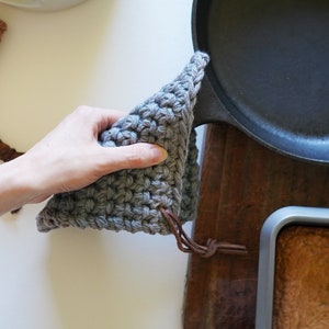 May include: Three crocheted pot holders in brown, green, and grey with leather straps. A hand is holding the grey pot holder over a black cast iron skillet.