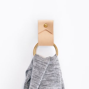 May include: A light-colored leather towel holder with a brass ring and a gray towel hanging from it. The holder is mounted on a white wall. A green plant is visible in the lower left corner.
