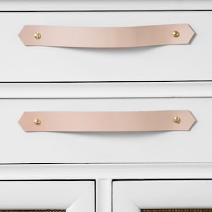 May include: White cabinet with two drawers and two doors. The drawers have light pink leather pulls with brass accents. The doors have a woven brown texture and light pink leather pulls with brass accents.
