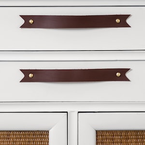 Keyaiira Chocolate genuine leather drawer handle with V-shaped pointed ends and polished brass mounting hardware installed horizontally on a white cabinet above woven cane doors, highlighting the rich Chocolate leather tone and handcrafted raised grip